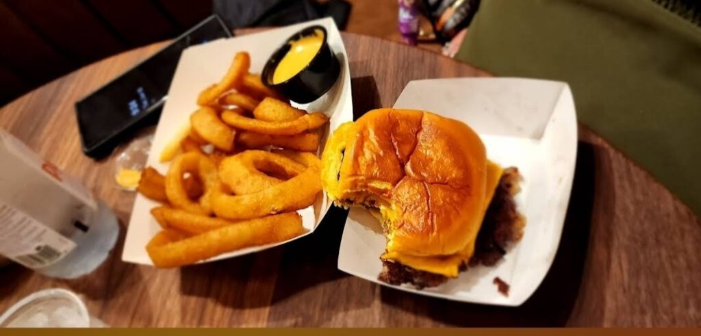 A juicy cheeseburger with a golden-brown bun, partially eaten, served on a white paper tray next to a side of crispy onion rings with a small cup of dipping sauce