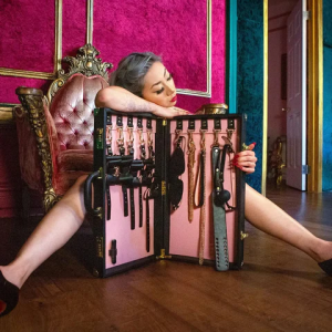 A woman sitting on the floor beside an open pink case filled with whips, paddles, and restraints, showcasing an elegant collection of BDSM Gear for beginners.